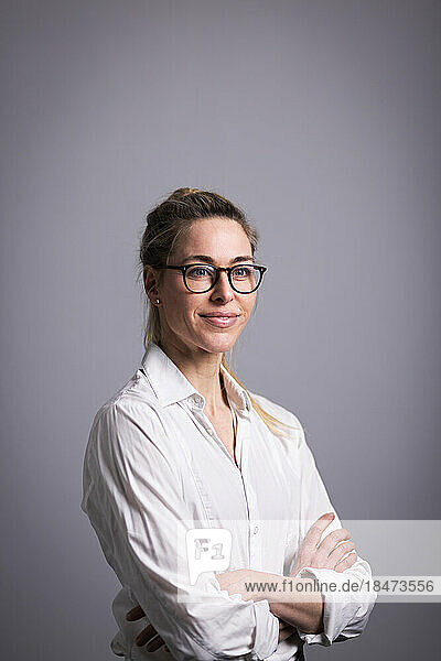 Smiling businesswoman wearing eyeglasses with arms crossed standing against gray background