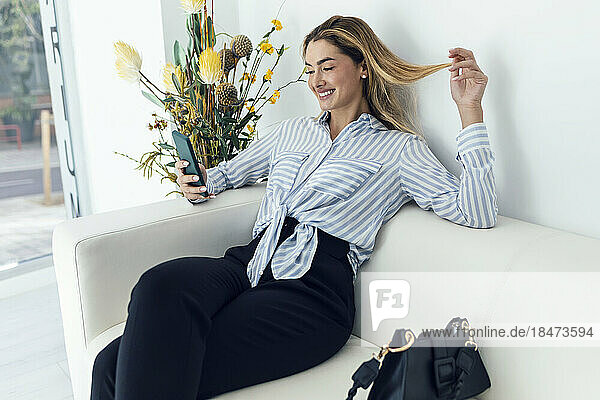 Lächelnde junge Frau  die auf dem Sofa sitzt und ihr Smartphone benutzt
