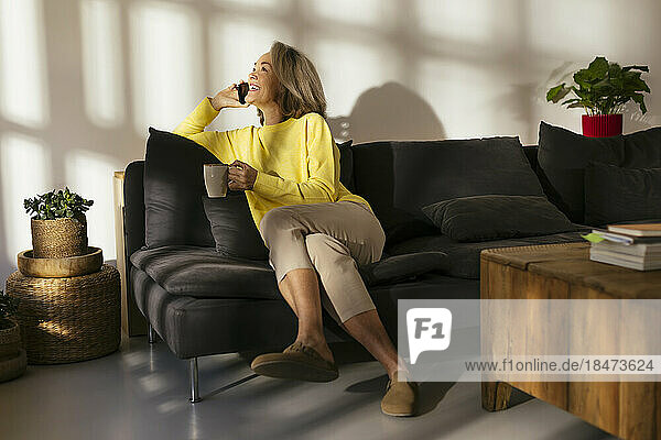 Happy woman with coffee cup talking on smart phone at home