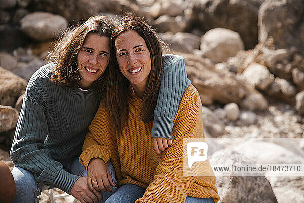 Smiling woman with arm around friend sitting on rocks