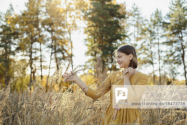 Smiling woman touching dry grass standing in field