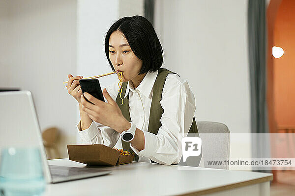 Businesswoman eating noodles and using smart phone in office