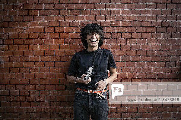 Happy young man standing with camera in front of brick wall