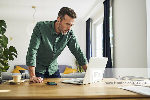 Reifer Mann lehnt am Schreibtisch und schaut auf den Laptop