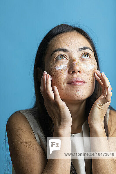 Woman applying cream on face against colored background
