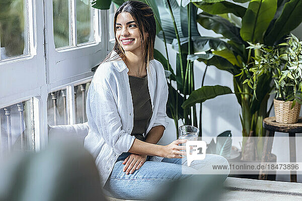 Happy young woman sitting with glass of water at the window