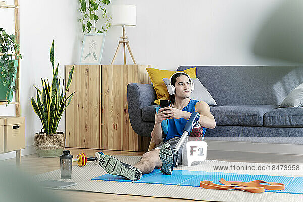 Thoughtful man with prosthetic leg holding smart phone in living room
