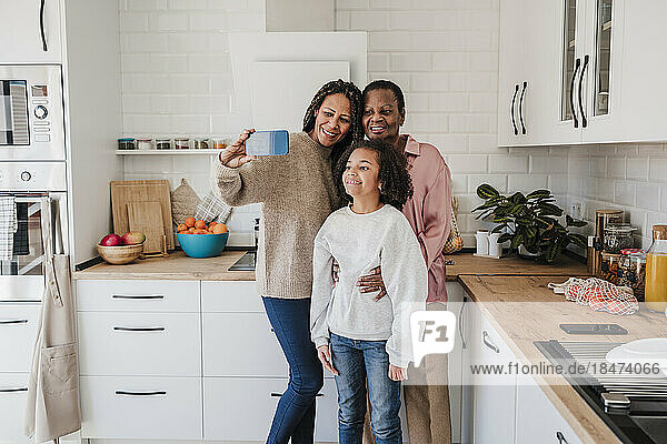 Woman taking selfie with mother and daughter through smart phone in kitchen at home