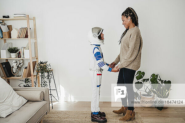 Smiling woman standing with daughter wearing space suit near wall at home