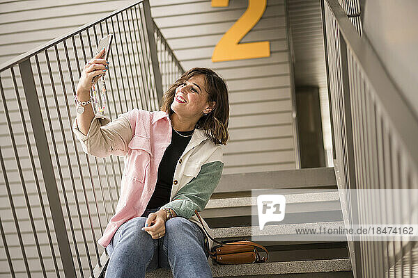 Happy woman taking selfie through smart phone sitting on stairs