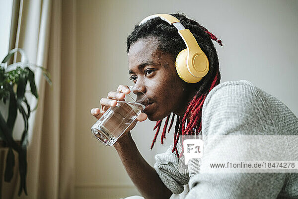 Nachdenklicher junger Mann mit Kopfhörern trinkt zu Hause Wasser