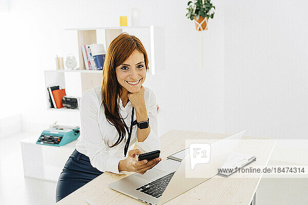Smiling female freelancer with mobile phone and laptop sitting at desk