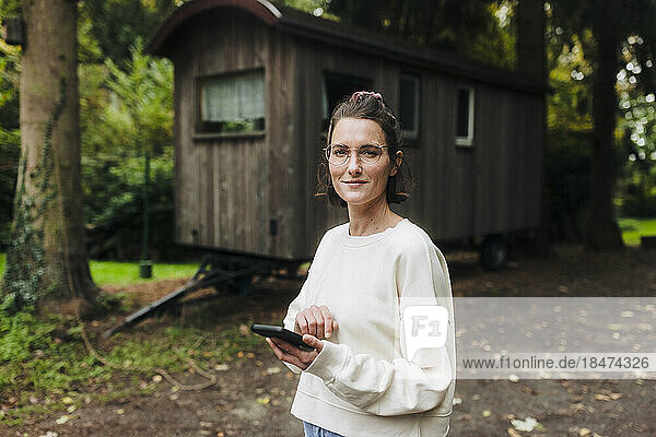 Smiling woman with smart phone standing in front of cabin