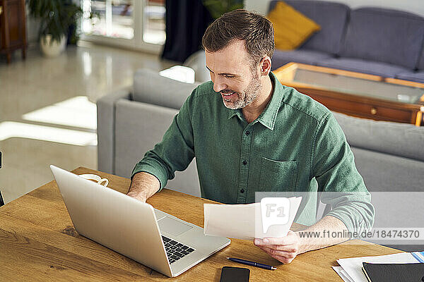 Smiling man working from home using laptop holding documents