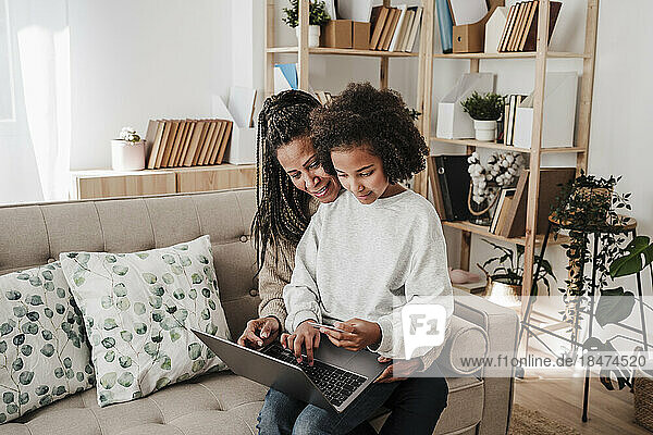 Tochter macht Online-Einkäufe mit Mutter über Laptop  die zu Hause auf dem Sofa sitzt
