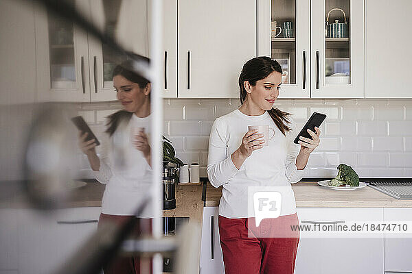 Lächelnde Frau benutzt Smartphone und Kaffeetasse in der Küche