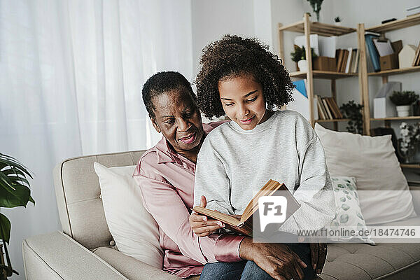Enkelin liest Buch mit Großmutter  die zu Hause auf dem Sofa sitzt