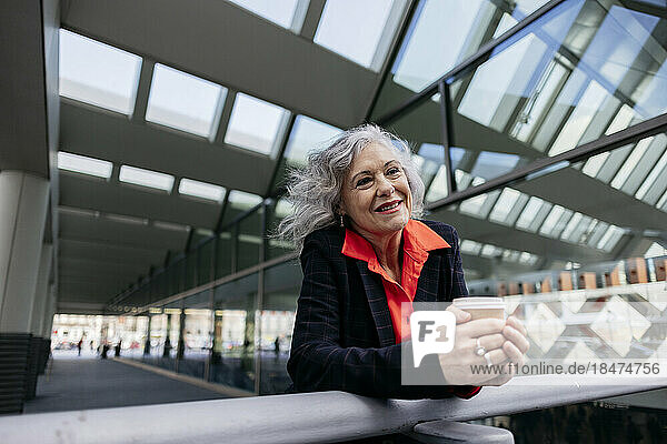Smiling mature businesswoman with disposable coffee cup leaning on railing