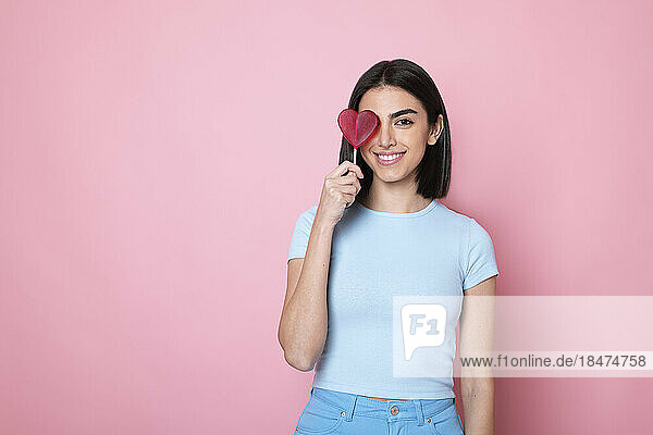 Happy woman holding heart shaped lollipop over eye against pink background