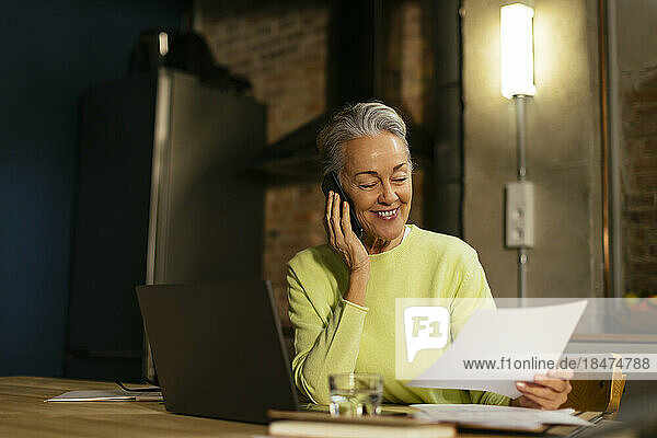 Happy freelancer holding document and talking on smart phone at desk