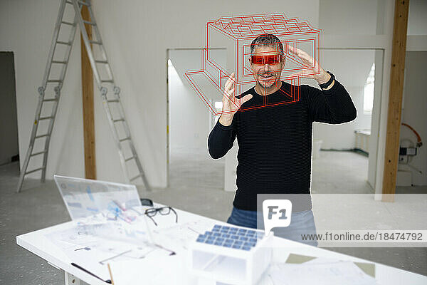 Smiling mature architect wearing virtual reality simulator examining house model at construction site