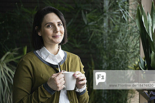 Smiling woman standing with coffee cup in front of plants