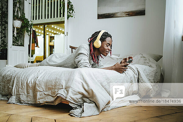 Smiling young man wearing headphones and using smart phone lying on bed at home