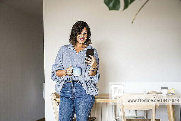 Glückliche Frau mit Kaffeetasse und Smartphone zu Hause