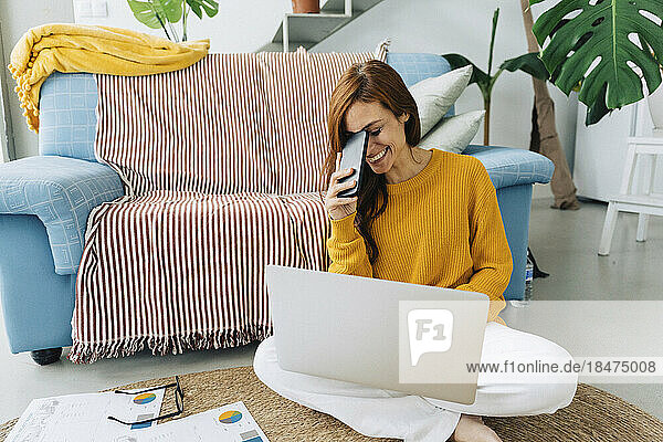 Happy woman with smart phone and laptop sitting on carpet at home