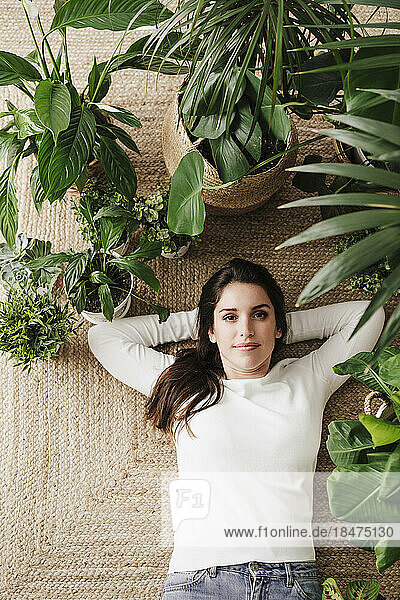 Woman with hands behind head lying amidst plants on carpet