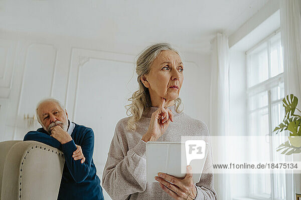 Besorgte ältere Frau hält Tablet-PC mit Mann im Hintergrund zu Hause