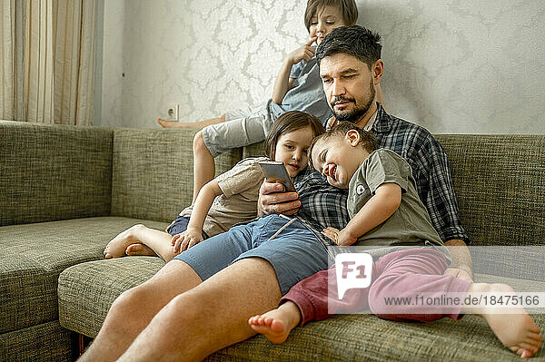 Father sharing smart phone with sons sitting on sofa at home