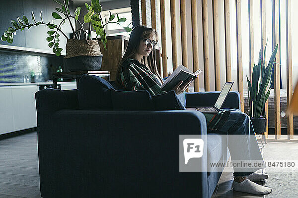 Young woman sitting with laptop reading book on sofa at home