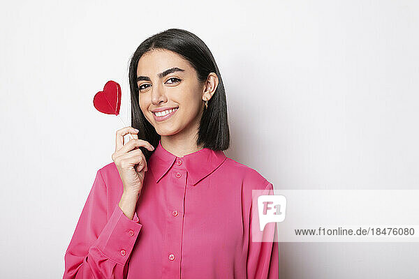 Happy young woman holding red heart shaped lollipop against white background