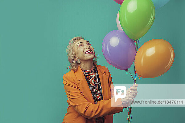 Happy senior woman holding colorful balloons against turquoise background