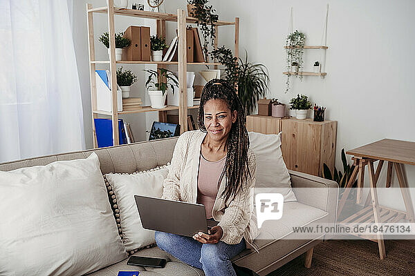 Smiling freelancer with laptop sitting on sofa at home office