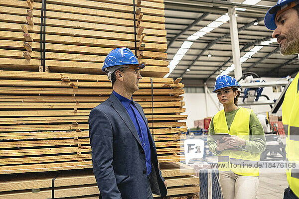 Mature engineer discussing with colleagues by stack of planks in factory