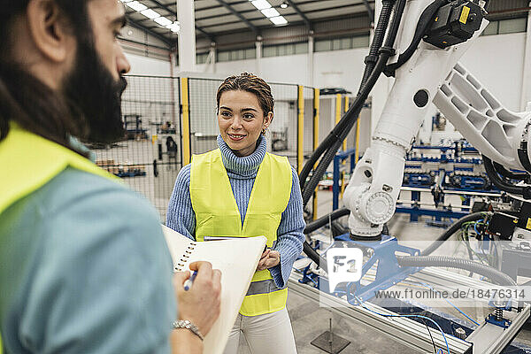 Smiling engineer looking at colleague with note pad standing by robotic arm