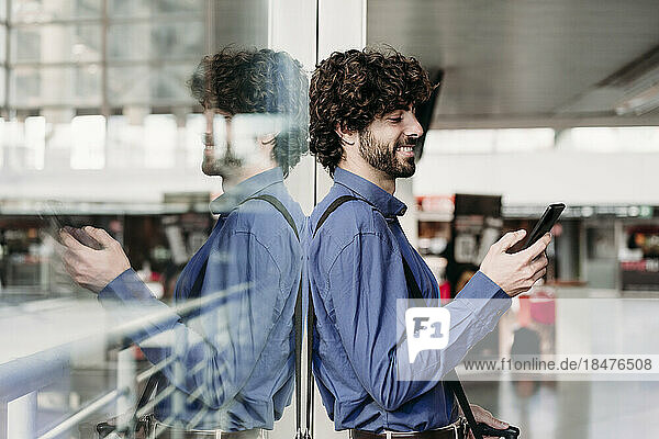 Lächelnder Geschäftsmann lehnt an Glas und benutzt Smartphone am Bahnhof