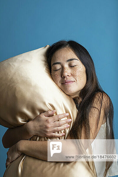 Frau mit geschlossenen Augen umarmt goldenes Kissen vor blauem Hintergrund