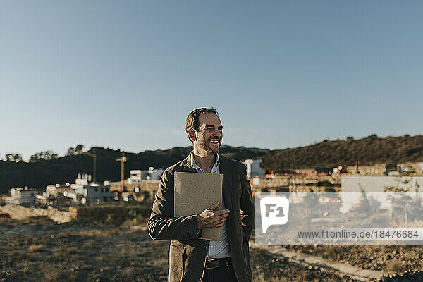 Lächelnder Mann hält Akte in der Hand und steht auf leerer Fläche für den Wohnungsbau