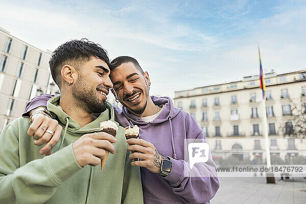 Happy gay couple with arms around holding ice cream at city street