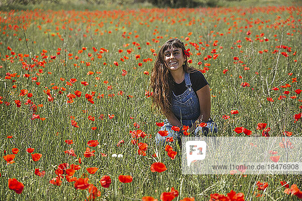 Happy young woman sitting amidst field with poppy flowers