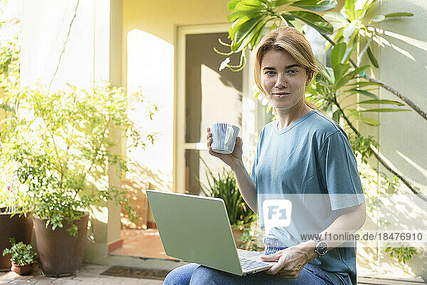Smiling freelancer with coffee cup and laptop sitting in garden