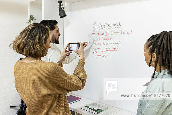 Businesswoman clicking photo of colleague explaining on white board
