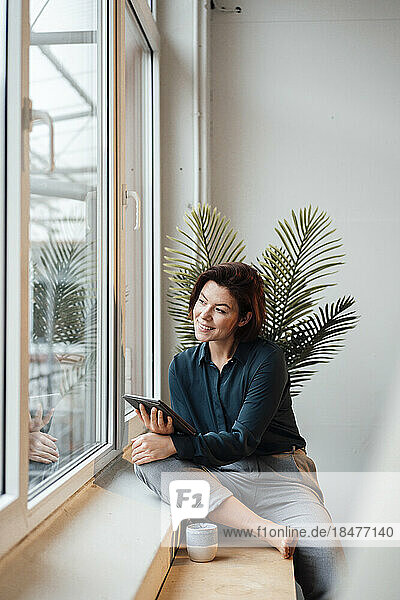 Smiling young businesswoman with tablet PC looking through window in office