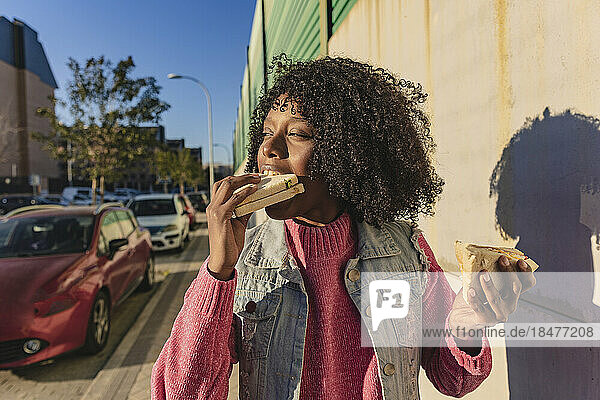Junge Frau isst an einem sonnigen Tag Sandwich am Fußweg