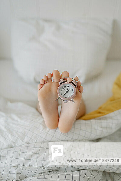Feet of girl with alarm clock on bed at home