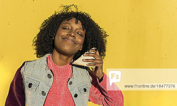 Smiling woman with sandwich standing in front of yellow wall