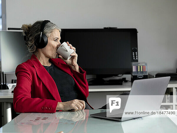 Senior businesswoman wearing wireless headphones drinking coffee in office
